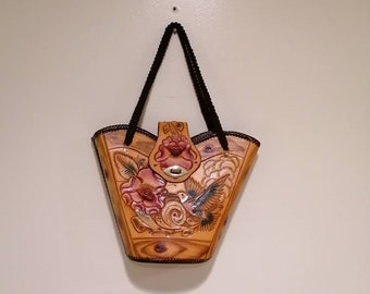 Peut inclure: Un sac fourre-tout en cuir beige avec un motif floral et un aigle. Le sac est doté d'une poignée noire tressée et d'un fermoir décoratif. Le motif floral comprend des roses aux accents roses et rouges.