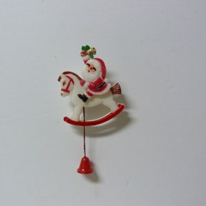 Op de afbeelding: Een wit schommelpaard met een rode basis en een kerstmanfiguur erop. De kerstmanfiguur draagt een rood pak met witte versieringen en een witte baard. Hij houdt een groene tak van hulst in zijn rechterhand. Het schommelpaard heeft een rode bel die aan de onderkant hangt.