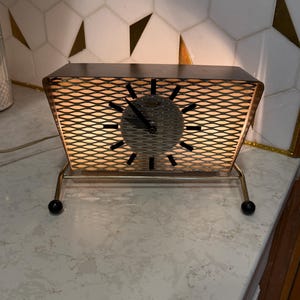 May include: Retro electric clock with a dark brown frame and a mesh front. The clock face has black hands and markers. It sits on a gold-coloured metal stand with black ball feet. The clock is lit from within.