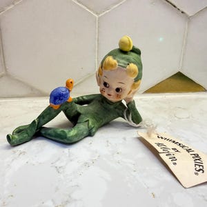 Peut inclure: Figurine en céramique d'un lutin en tenue verte, allongé avec une petite tortue. Le lutin a les cheveux blonds et un chapeau jaune. La tortue est bleue et orange. Une étiquette indique "Whimsical Pixies by Hellerin."