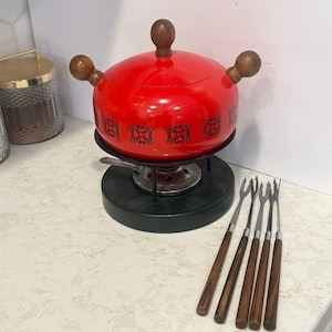 May include: A bright red fondue pot with a lid, featuring dark wooden handles and a black decorative pattern around the base. The pot sits on a black metal stand with a burner and is accompanied by five metal fondue forks with dark wooden handles.