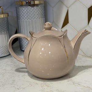 May include: A pale pink ceramic teapot with a floral design and gold detailing. The teapot features a curved handle and a lid with a bud-shaped knob. It sits on a white countertop, with a decorative tile backsplash in the background.