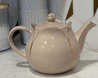 Può includere: Una teiera in ceramica rosa pallido con un design floreale e finiture dorate. La teiera ha un manico curvo e un coperchio con un pomello a forma di bocciolo. È appoggiata su un piano di lavoro bianco, con un paraschizzi in piastrelle decorative.