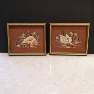 May include: Two framed embroidered seashell art pieces. Each features a detailed seashell design with various colors, including white, beige, and brown, set against a rust-colored background. The frames are a light gold color.