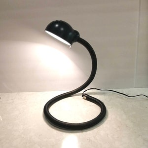 Peut inclure: Une lampe de bureau à col flexible noire avec un abat-jour rond noir. La lampe est allumée et éclaire la zone autour d'elle.
