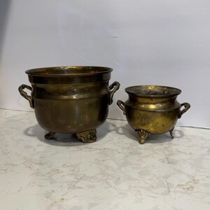 May include: Two brass pots with a weathered patina, each featuring two side handles and three decorative feet. The larger pot is positioned on the left, while the smaller pot sits to the right, both displaying a rounded, metallic body.