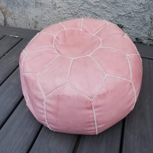 Op de afbeelding: Een roze leren poef met witte stiksels. De poef heeft een ronde vorm en is gemaakt van zacht, soepel leer.