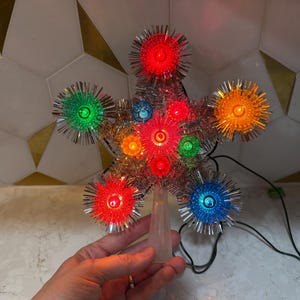 May include: A vintage Christmas tree topper shaped like a star. It has colourful lights in red, green, orange, and blue, surrounded by silver tinsel. The base is made of clear plastic.