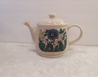 May include: A white ceramic teapot with a speckled glaze and a floral design featuring blue, pink, and yellow flowers. The teapot has a spout and a handle.