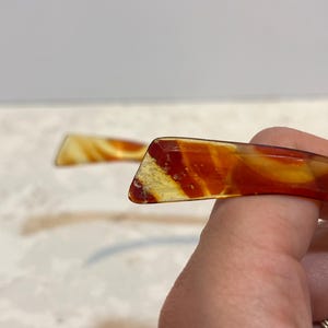 May include: Two vintage hair combs with a marbled pattern in shades of amber, yellow, and red. The combs have a rectangular shape with a slightly curved design. One comb is in focus, showing the details of the material.