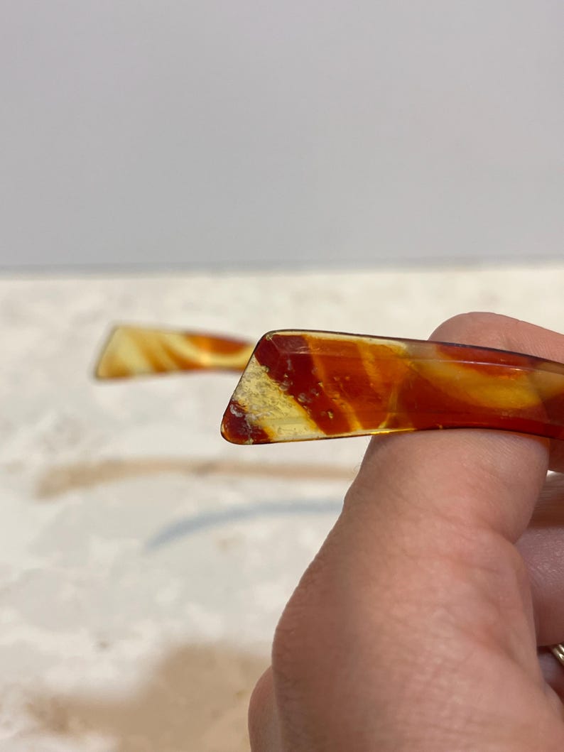 May include: Two vintage hair combs with a marbled pattern in shades of amber, yellow, and red. The combs have a rectangular shape with a slightly curved design. One comb is in focus, showing the details of the material.