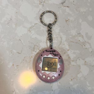 May include: A pink Tamagotchi Angel keychain with a silver chain and ring. The digital pet device displays a pixelated character on its screen. The text "Tamagotchi Angel" is above the screen, with star and wing details.