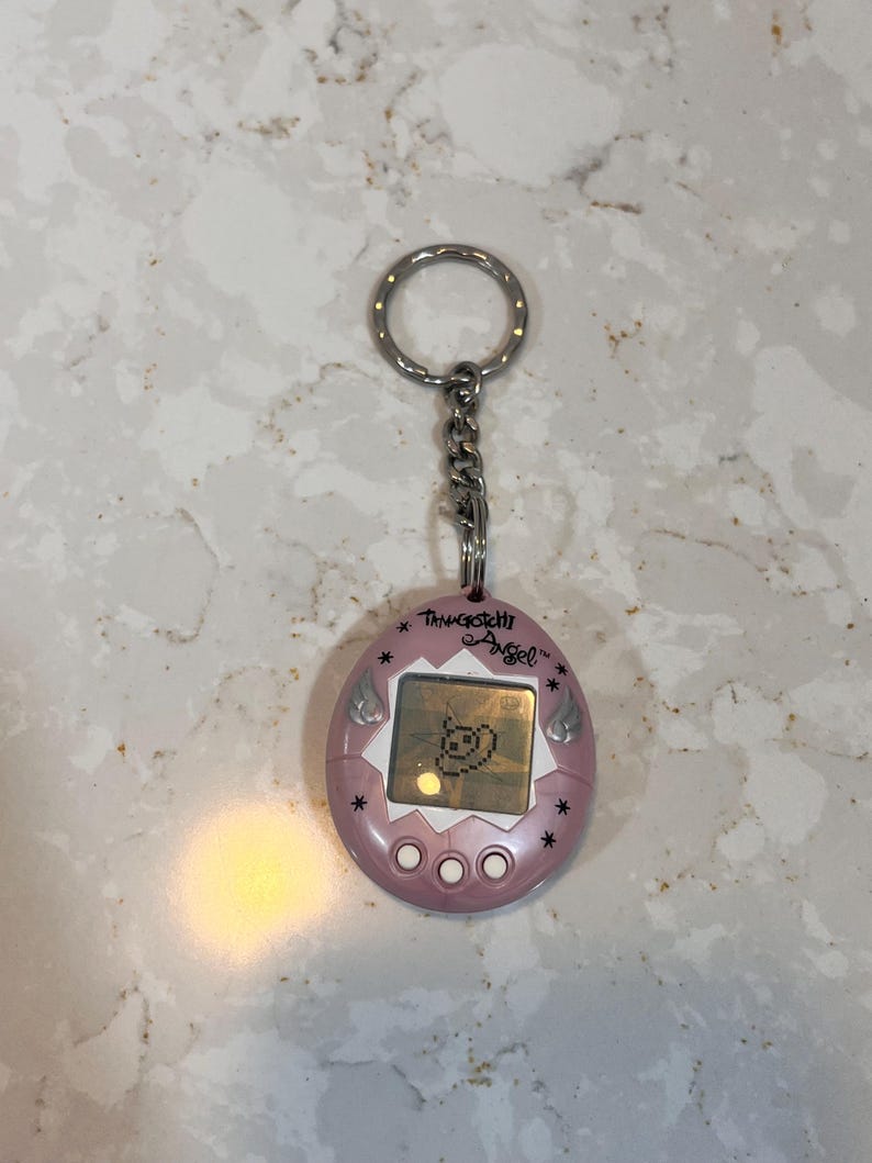 May include: A pink Tamagotchi Angel keychain with a silver chain and ring. The digital pet device displays a pixelated character on its screen. The text "Tamagotchi Angel" is above the screen, with star and wing details.