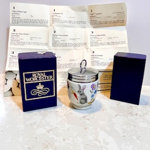 May include: A white porcelain egg cup with a silver lid and a floral design featuring a rabbit. The egg cup is sitting on a white countertop. The box for the egg cup is blue with gold lettering that reads "Royal Worcester".