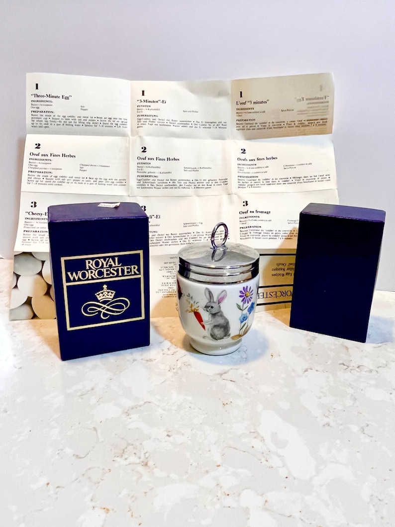 May include: A white porcelain egg cup with a silver lid and a floral design featuring a rabbit. The egg cup is sitting on a white countertop. The box for the egg cup is blue with gold lettering that reads "Royal Worcester".