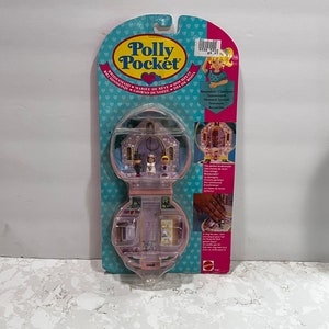 Op de afbeelding: Een roze Polly Pocket speelgoed met een kerkscène erin. De kerk heeft een bruidspaar erin en een ringendoos aan de zijkant. Het speelgoed is verpakt in een doorzichtige plastic clamshell met een blauwe en roze achtergrond. De tekst op de verpakking luidt "Polly Pocket" en "Keepsakes Classiques".