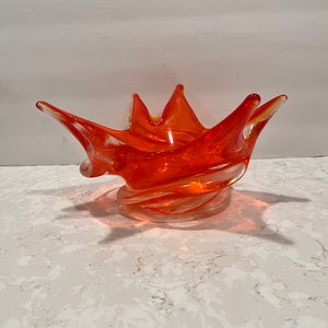 May include: Orange glass bowl with a unique, spiky, and swirled design. The bowl is made of clear glass with an orange tint.