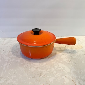 May include: Orange enamel cast iron pot with a black knob on the lid and a single handle. The pot is sitting on a white countertop.