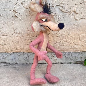 Pode incluir: Um brinquedo de pelúcia vintage de um coiote de desenho animado, principalmente rosa com um peito e rosto creme. O coiote tem um focinho longo, um nariz preto e cabelo preto. O brinquedo está em uma superfície de concreto.