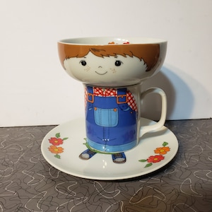 May include: A ceramic mug and saucer set featuring a cartoon character design. The mug depicts a figure in blue overalls and a red checkered shirt, while the bowl-shaped top shows a face with brown hair. The white saucer features orange and red flowers.