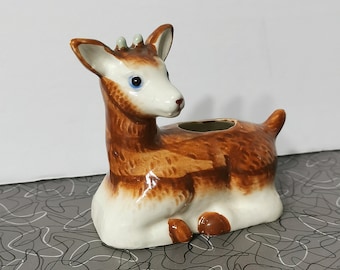 May include: A ceramic deer planter with brown and white markings. The deer is lying down with its head turned to the side. The planter has a small opening on the top for planting.
