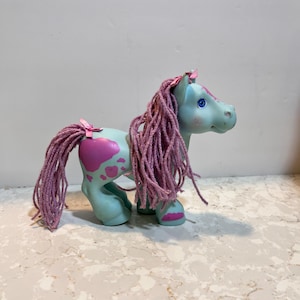 Könnte beinhalten: Ein Vintage-Spielzeugpony in Hellblau mit rosa Mähne und Schweif. Das Pony hat herzförmige rosa Markierungen an der Seite und eine rosa Schleife am Schwanz. Die Augen des Ponys sind blau.