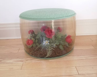 May include: A clear plastic ottoman with a green top and a floral arrangement inside. The arrangement includes red roses and white flowers.