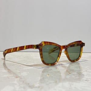 May include: Vintage sunglasses with a tortoiseshell pattern in shades of brown and yellow. The frames have a unique shape, and the lenses are a muted green color. The sunglasses are resting on a reflective surface.