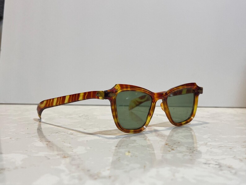May include: Vintage sunglasses with a tortoiseshell pattern in shades of brown and yellow. The frames have a unique shape, and the lenses are a muted green color. The sunglasses are resting on a reflective surface.