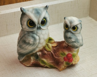 May include: Two ceramic owls perched on a tree branch with green leaves and red berries. The larger owl has yellow eyes and a gray and white patterned body. The smaller owl has yellow eyes and a white body.
