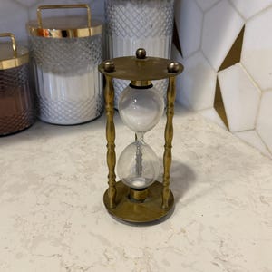 Könnte beinhalten: Eine antike Sanduhr aus Messing mit durchsichtigen Glaskolben. Die Sanduhr hat einen runden Boden und eine runde Oberseite, die durch vier verzierte Messingstützen verbunden sind. Der Sand im Inneren ist weiß. Dekorative Gläser mit goldenen Deckeln sind im Hintergrund.