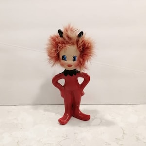 Peut inclure: Une figurine de diable rouge avec des cornes noires et des cheveux roux et jaunes duveteux. La figurine a des yeux bleus et une bouche noire.