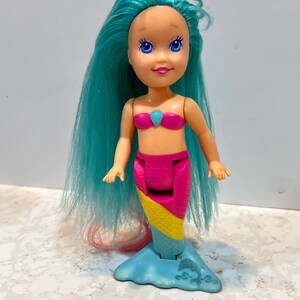 May include: A plastic mermaid doll with long, turquoise hair and a pink top. The doll has a pink and yellow tail, and a blue base. The mermaid's face has blue eyes and a smiling expression.