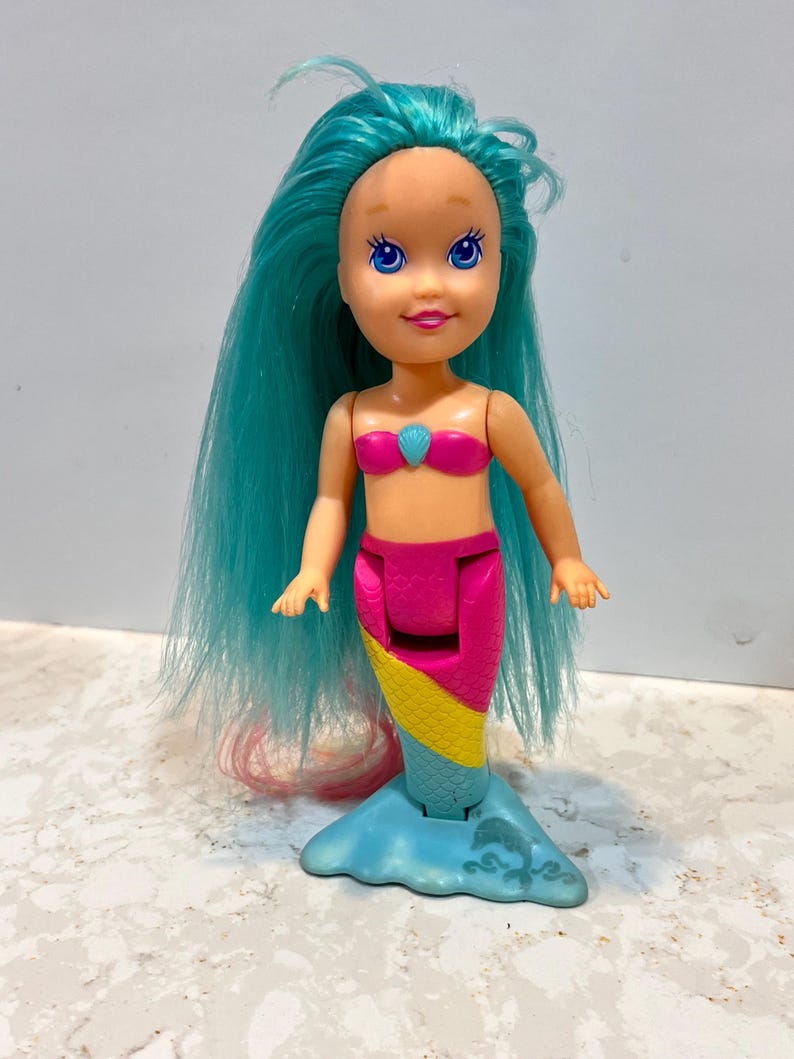 May include: A plastic mermaid doll with long, turquoise hair and a pink top. The doll has a pink and yellow tail, and a blue base. The mermaid's face has blue eyes and a smiling expression.