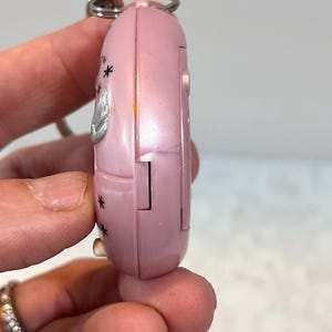 May include: A pink, handheld electronic device with a silver keyring attached. The device has a rounded shape with a hinged section and is decorated with small black star shapes and a silver emblem.