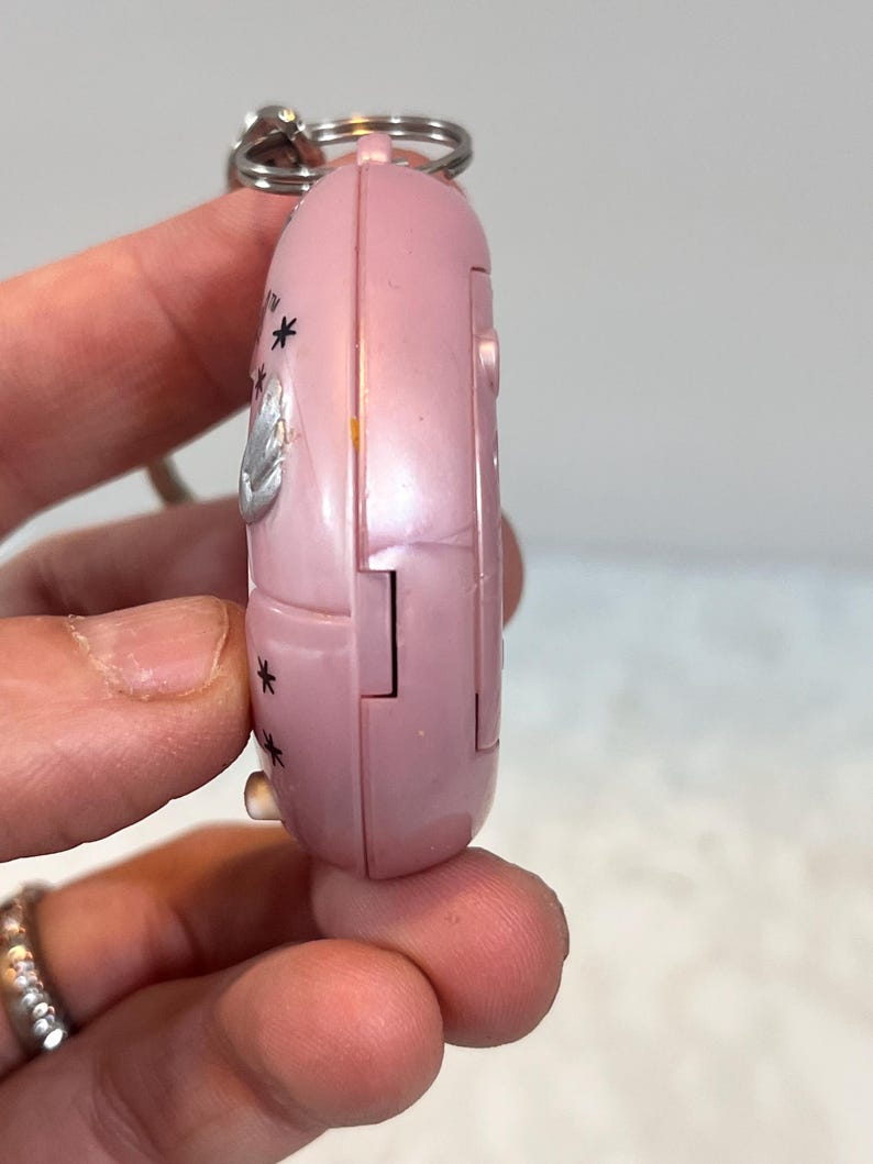 May include: A pink, handheld electronic device with a silver keyring attached. The device has a rounded shape with a hinged section and is decorated with small black star shapes and a silver emblem.