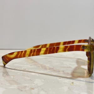 May include: Side view of eyeglasses with a tortoiseshell pattern. The frames are a mix of yellow and brown, with a rectangular shape. The temples have a striped design. The glasses are set against a white background.