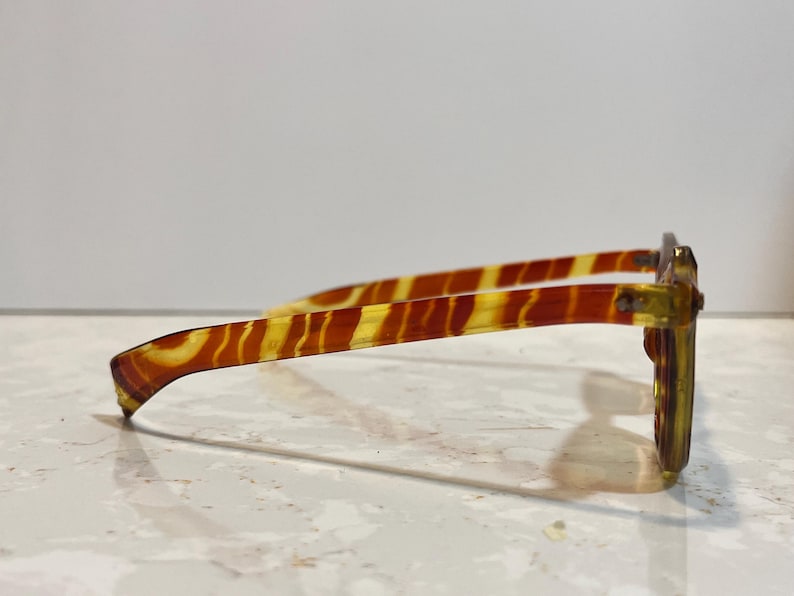 May include: Side view of eyeglasses with a tortoiseshell pattern. The frames are a mix of yellow and brown, with a rectangular shape. The temples have a striped design. The glasses are set against a white background.