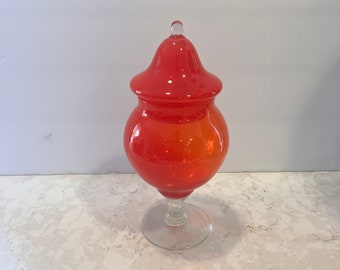 May include: A red glass jar with a lid and a clear glass base. The jar has a rounded body and a flared top. The lid is domed and has a small knob on top.