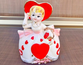 May include: A white ceramic figurine of a girl wearing a red heart-shaped hat and a pink and white dress with red hearts. She is holding a basket of red hearts.