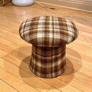 Könnte beinhalten: Ein pilzförmiger Hocker mit einem braunen und cremefarbenen Karomuster. Der Hocker hat eine runde Oberseite und einen ausgestellten Sockel. Der Stoff scheint ein strukturiertes Gewebe zu sein. Es ist ein dekoratives Möbelstück.