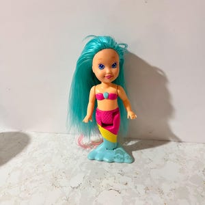 May include: A small mermaid doll with blue hair, a pink seashell top, and a colorful tail. The tail transitions from pink to yellow to blue. The doll has a smiling face and is standing on a light blue base.