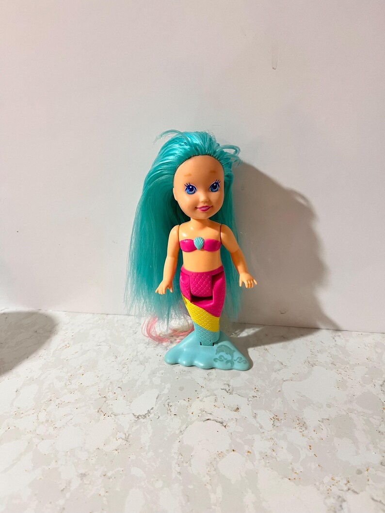 May include: A small mermaid doll with blue hair, a pink seashell top, and a colorful tail. The tail transitions from pink to yellow to blue. The doll has a smiling face and is standing on a light blue base.