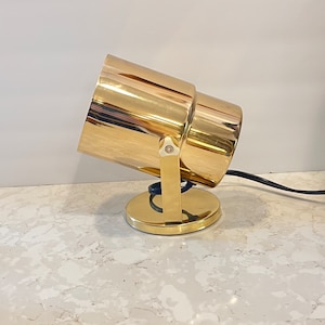 Puede incluir: Un foco de luz dorado con una base redonda y una cabeza giratoria. La luz está sobre una superficie de mármol blanco.