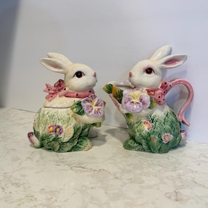 May include: A ceramic sugar bowl and creamer set shaped like white rabbits. Each rabbit features a pink polka-dot bow, green textured foliage, and purple pansy flower accents. The creamer has a curved handle and a spout, while the sugar bowl has a lid.
