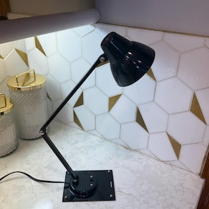 May include: Black adjustable desk lamp with a rectangular base. The lamp has a black shade and a flexible arm. The lamp is on a white countertop with a decorative tile backsplash.