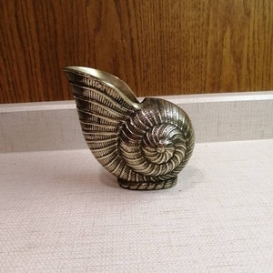 Könnte beinhalten: Ein metallisch goldfarbenes Messinggefäß in Form eines Nautilus-Muschelgehäuses. Das Objekt weist ein detailliertes Spiralmuster mit tiefen Rillen und Graten auf der Außenfläche auf, was ein strukturiertes Aussehen mit polierter Oberfläche erzeugt.