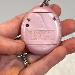 May include: A pink, handheld electronic pet toy with a silver keyring. The back of the toy has the text "BANDAI 1997" and "MADE IN CHINA". The toy is oval-shaped with a pearlescent finish and two visible screws.