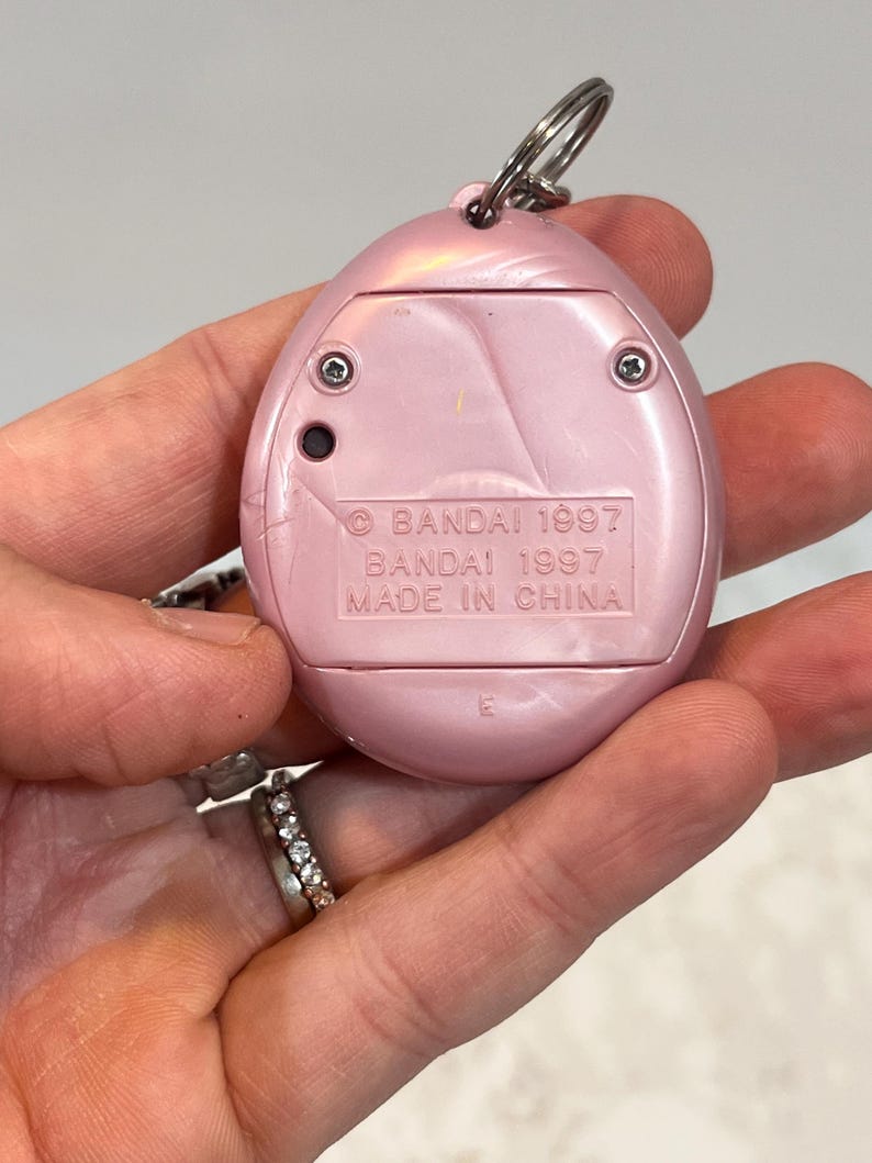 May include: A pink, handheld electronic pet toy with a silver keyring. The back of the toy has the text "BANDAI 1997" and "MADE IN CHINA". The toy is oval-shaped with a pearlescent finish and two visible screws.