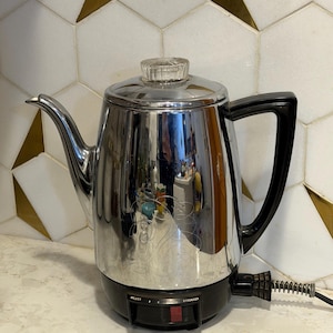 May include: A vintage chrome coffee percolator with a black base and handle. The percolator has a clear glass knob on the lid and a spout for pouring. The base has a dial with settings for "weaker" and "stronger".
